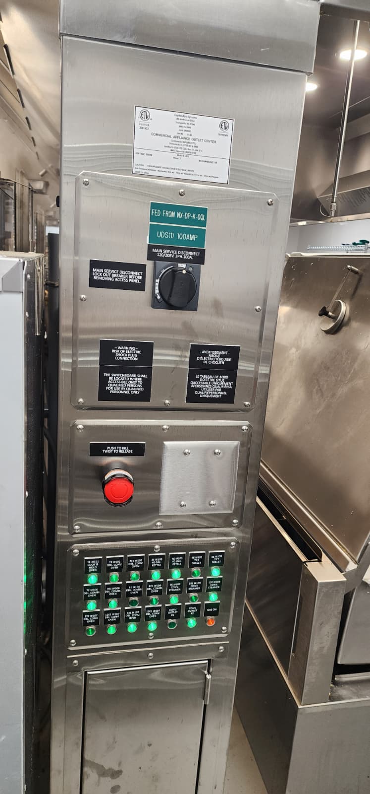 CaptiveAire Commercial Appliance Outlet Center stainless cabinet — Schneider Electric black main service disconnect handle in the OFF position with a yellow safety tag attached, below it a red mushroom-head emergency e-stop with a key cylinder and PUSH TO KILL / TWIST TO RELEASE label, both above the labeled grid of green LED appliance breakers